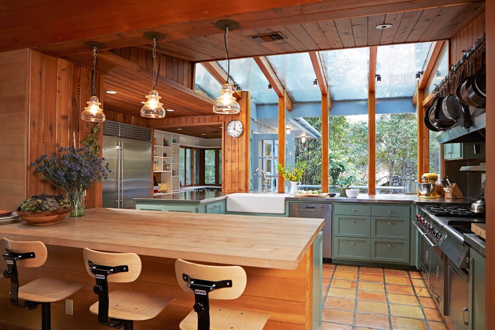 Photo of a country u-shaped kitchen in Los Angeles with terra-cotta floors, a farmhouse sink, shaker cabinets, green cabinets, stainless steel benchtops, stainless steel appliances, a peninsula and orange floor.