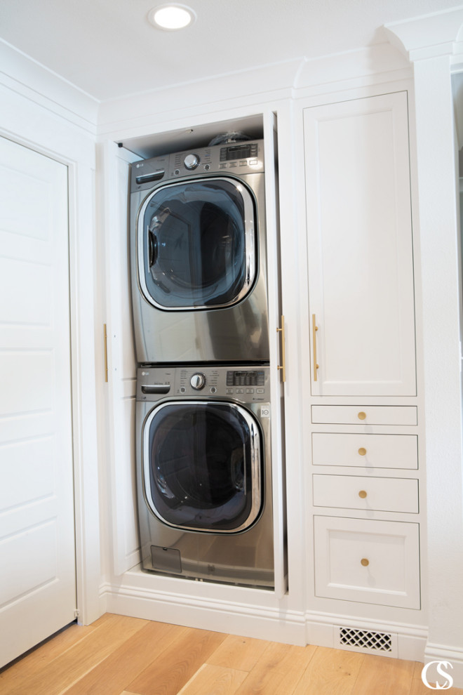 Modelo de armario lavadero tradicional con puertas de armario blancas, paredes blancas, suelo de madera clara y lavadora y secadora apiladas