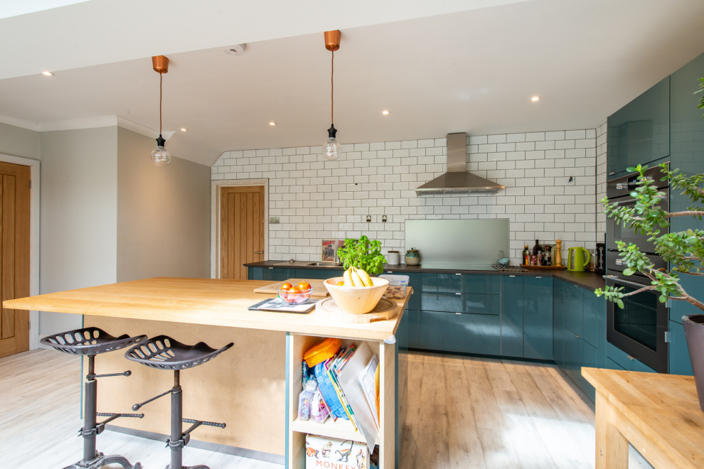 Photo of a contemporary l-shaped kitchen in Kent with flat-panel cabinets, blue cabinets, white splashback, subway tile splashback, stainless steel appliances, light hardwood floors, with island, beige floor and black benchtop.