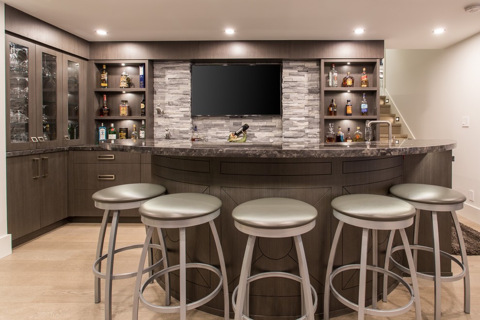 Large contemporary l-shaped seated home bar in Vancouver with an undermount sink, brown cabinets, marble benchtops, grey splashback, medium hardwood floors, beige floor, black benchtop, flat-panel cabinets and limestone splashback.