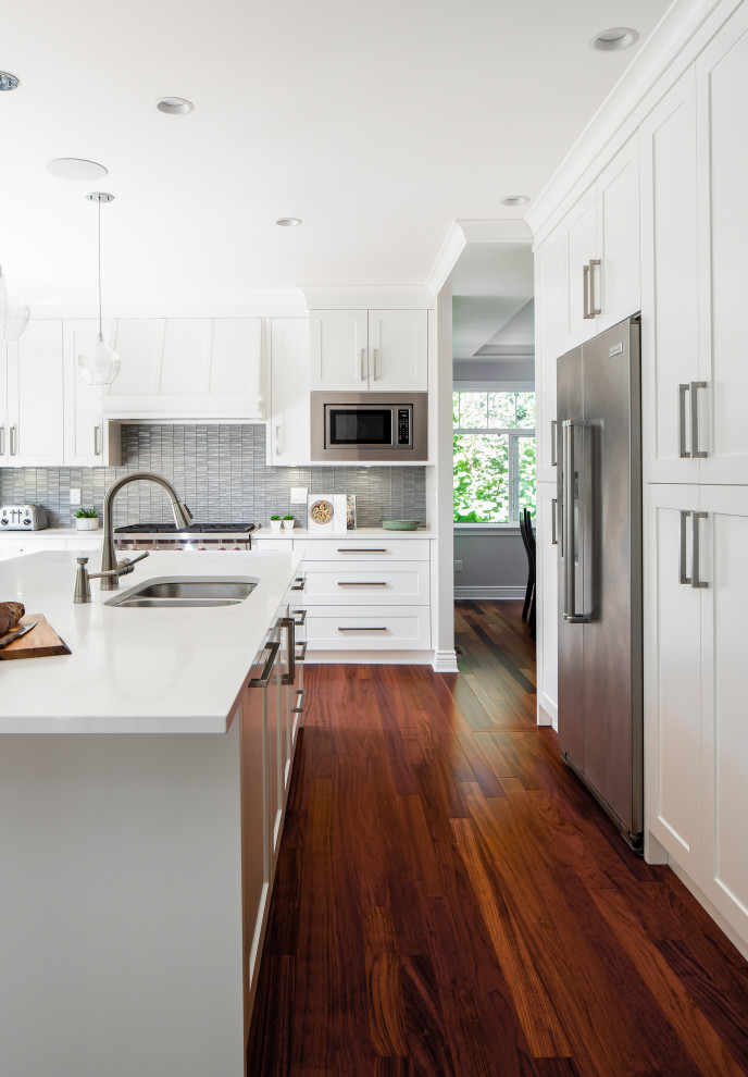 Design ideas for a transitional u-shaped kitchen in Vancouver with an undermount sink, shaker cabinets, white cabinets, grey splashback, mosaic tile splashback, stainless steel appliances, dark hardwood floors, with island, brown floor and white benchtop.