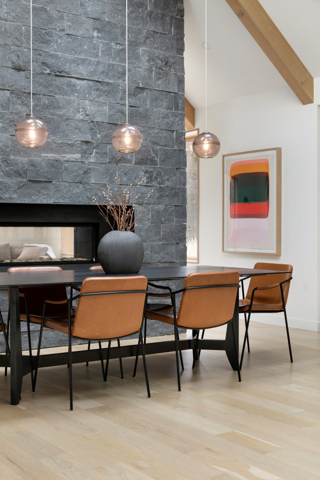 Photo of a contemporary kitchen/dining combo in Minneapolis with white walls, light hardwood floors, a two-sided fireplace, a stone fireplace surround, beige floor, exposed beam and vaulted.