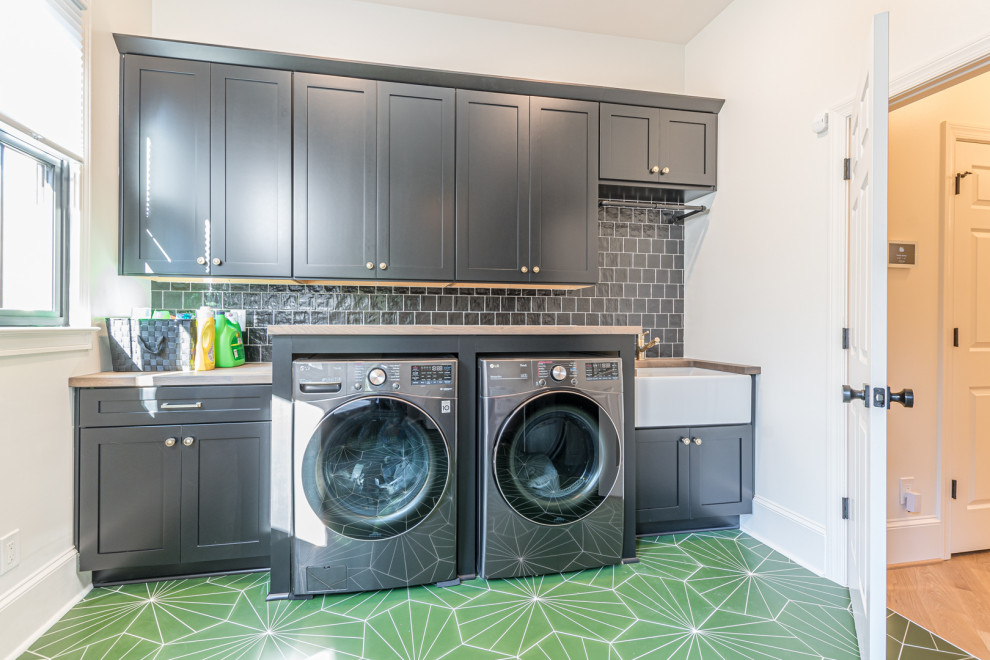 This is an example of a mid-sized transitional single-wall dedicated laundry room in Atlanta with a farmhouse sink, recessed-panel cabinets, black cabinets, wood benchtops, black splashback, stone tile splashback, white walls, a side-by-side washer and dryer, green floor and beige benchtop.