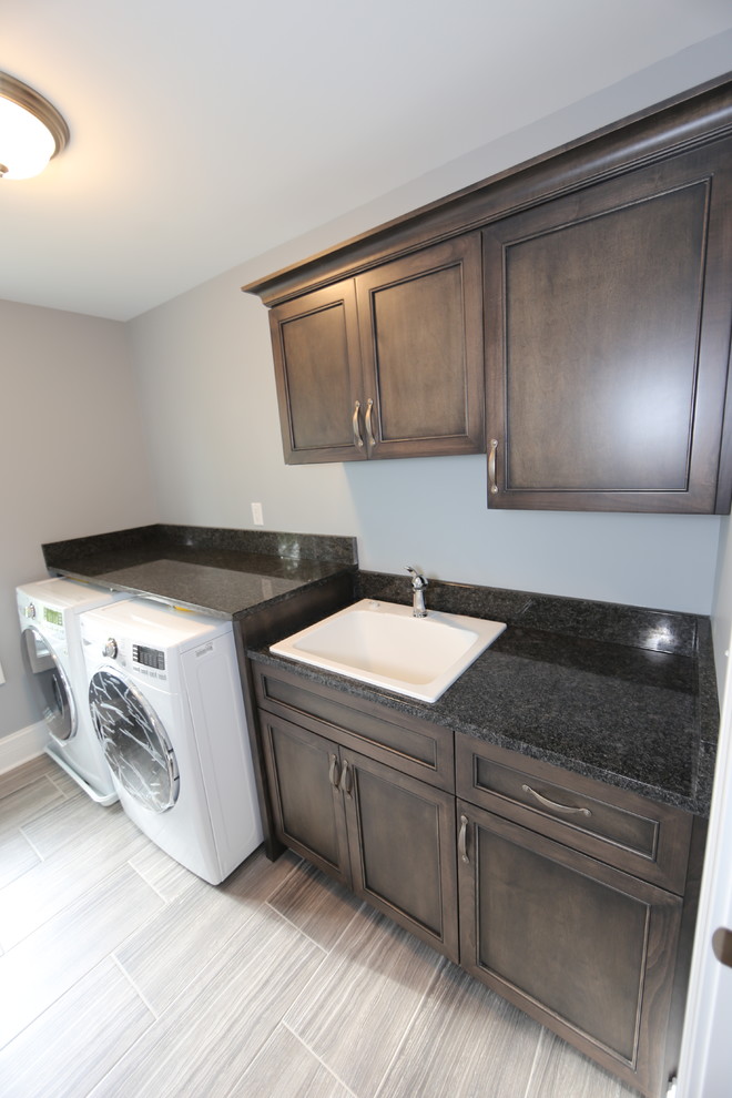 Mid-sized arts and crafts single-wall dedicated laundry room in Chicago with a drop-in sink, flat-panel cabinets, granite benchtops, grey walls, porcelain floors, a side-by-side washer and dryer, grey floor, black benchtop and dark wood cabinets.