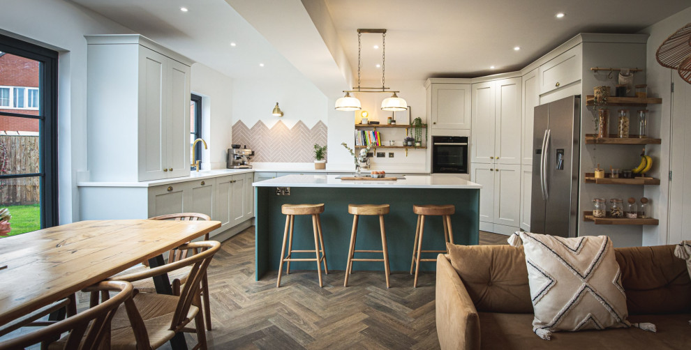 This is an example of a mid-sized traditional l-shaped open plan kitchen in Devon with an integrated sink, shaker cabinets, green cabinets, black appliances, with island, brown floor and white benchtop.