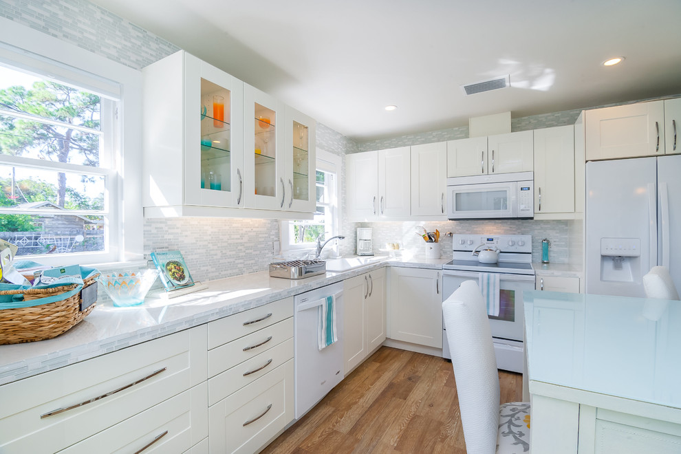 Photo of a small traditional l-shaped kitchen in Tampa with a single-bowl sink, shaker cabinets, white cabinets, multi-coloured splashback, glass sheet splashback, white appliances and light hardwood floors.