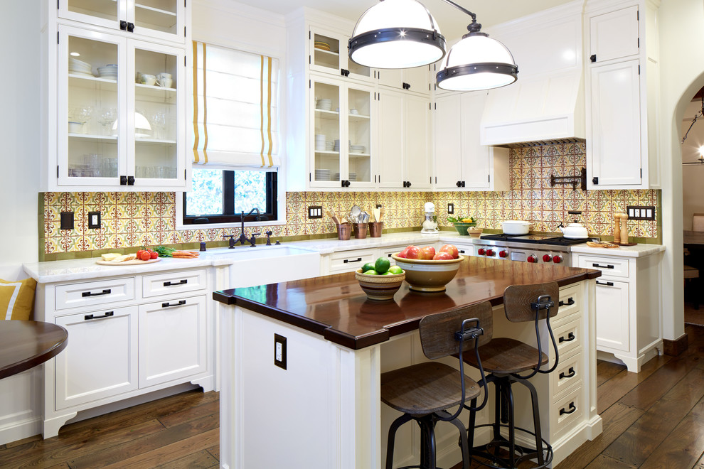 Mid-sized mediterranean l-shaped eat-in kitchen in Los Angeles with a farmhouse sink, glass-front cabinets, white cabinets, limestone benchtops, yellow splashback, mosaic tile splashback, with island, stainless steel appliances, dark hardwood floors and brown floor.