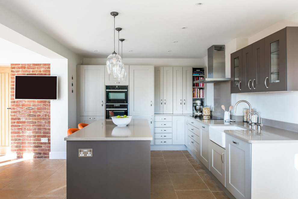 Mid-sized contemporary l-shaped open plan kitchen in Buckinghamshire with shaker cabinets, grey cabinets, with island, a farmhouse sink, grey splashback, black appliances, beige floor and grey benchtop.