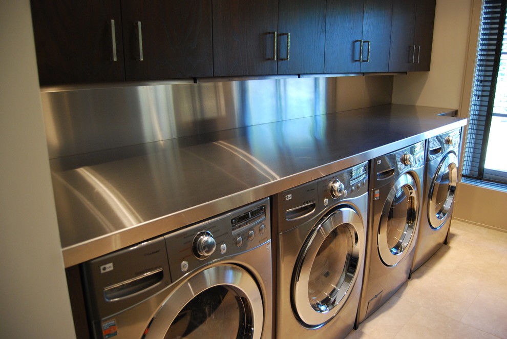 Photo of a mid-sized traditional single-wall dedicated laundry room in Chicago with flat-panel cabinets, dark wood cabinets, stainless steel benchtops, travertine floors, a side-by-side washer and dryer, white walls and beige floor.