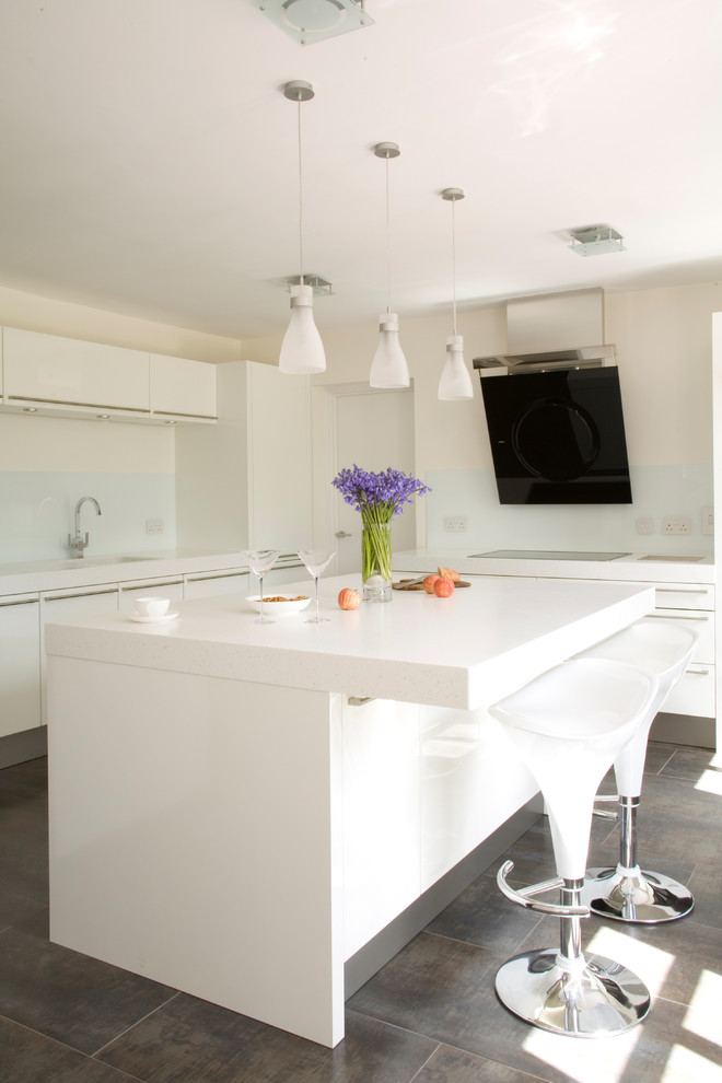 Large contemporary l-shaped eat-in kitchen in Sussex with a drop-in sink, flat-panel cabinets, white cabinets, solid surface benchtops, white splashback, glass sheet splashback, black appliances, porcelain floors, a peninsula, brown floor and white benchtop.
