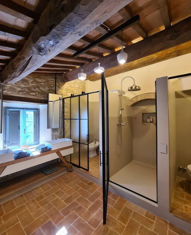 This is an example of a large traditional master bathroom in Florence with open cabinets, beige cabinets, a claw-foot tub, a curbless shower, a two-piece toilet, beige tile, limestone, beige walls, brick floors, a vessel sink, wood benchtops, brown floor, a hinged shower door, brown benchtops, a double vanity, a built-in vanity, exposed beam and brick walls.