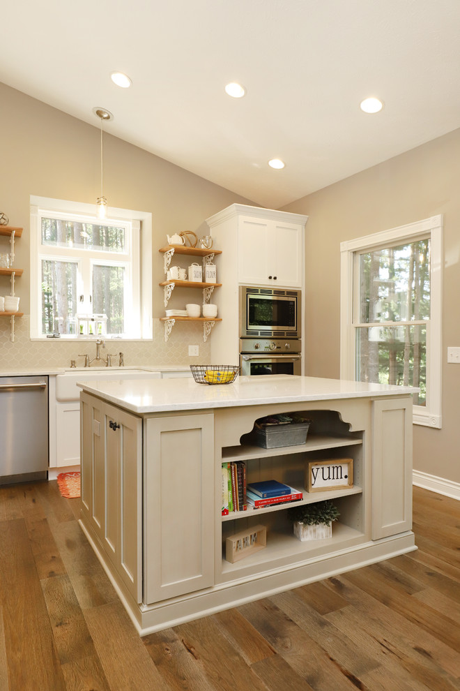 Design ideas for a mid-sized country l-shaped eat-in kitchen in Grand Rapids with a farmhouse sink, shaker cabinets, white cabinets, quartz benchtops, grey splashback, ceramic splashback, stainless steel appliances, light hardwood floors, with island and brown floor.