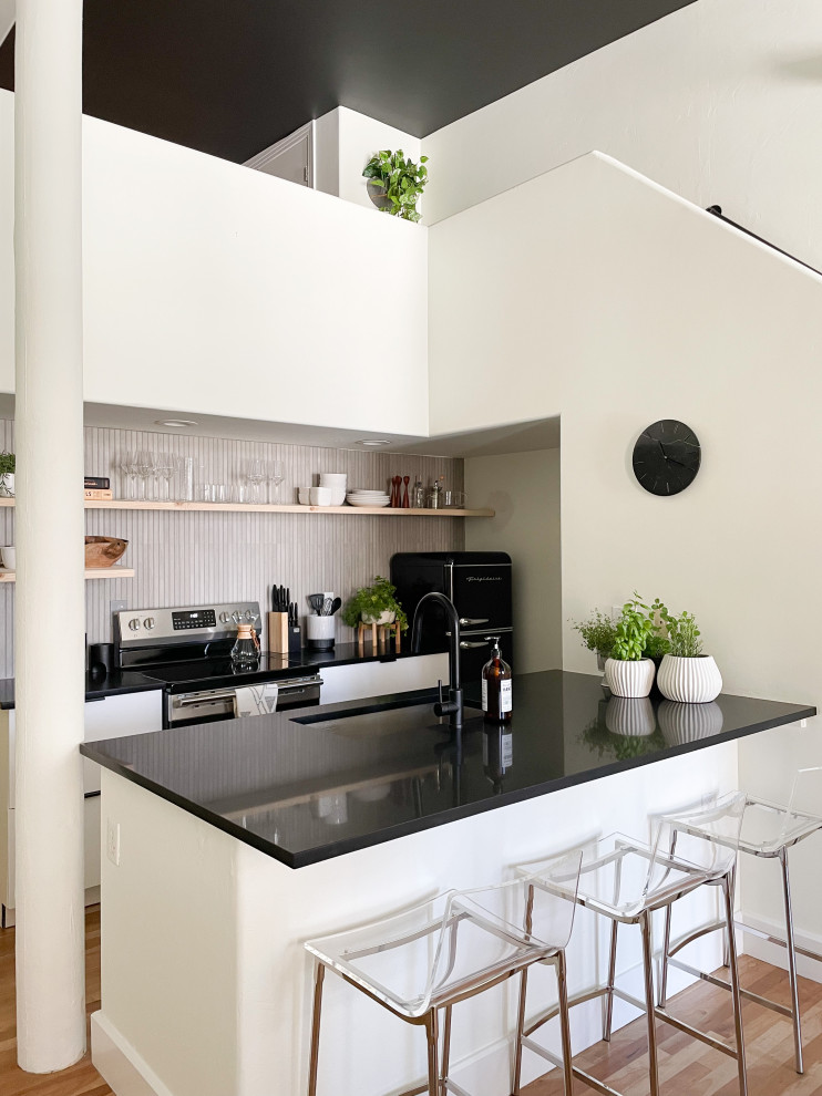 Small industrial galley open plan kitchen in Denver with an undermount sink, open cabinets, solid surface benchtops, grey splashback, cement tile splashback, black appliances, light hardwood floors, black benchtop and a peninsula.