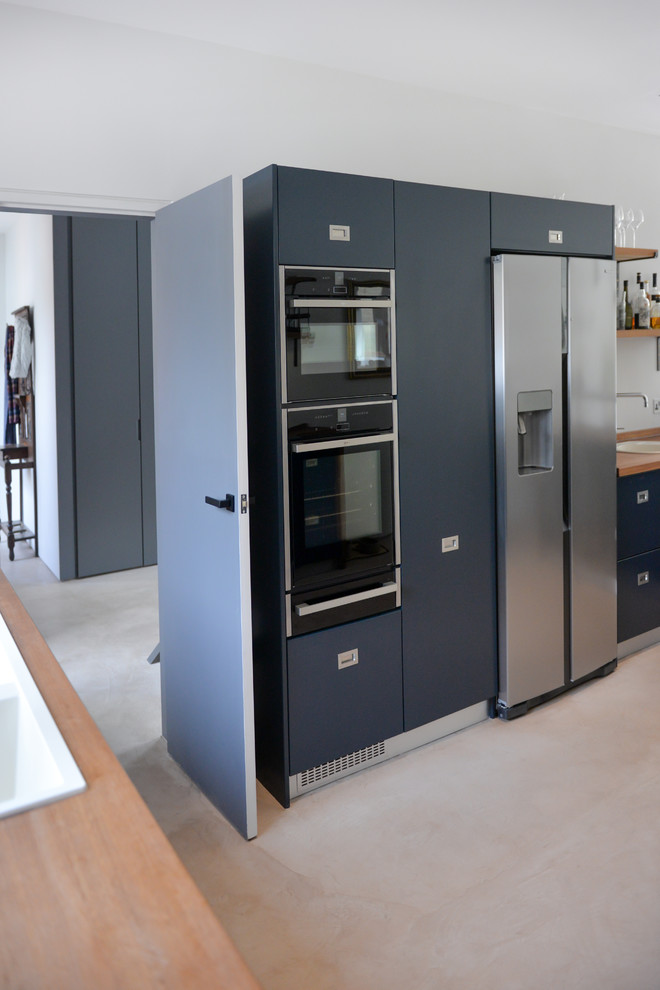 Photo of a mid-sized contemporary open plan kitchen in Sussex with a drop-in sink, flat-panel cabinets, blue cabinets, wood benchtops, black splashback, ceramic splashback, black appliances, concrete floors and with island.