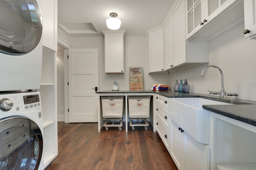 Inspiration for a large transitional l-shaped dedicated laundry room in Minneapolis with a farmhouse sink, recessed-panel cabinets, white cabinets, soapstone benchtops, grey walls, dark hardwood floors, a stacked washer and dryer and brown floor.