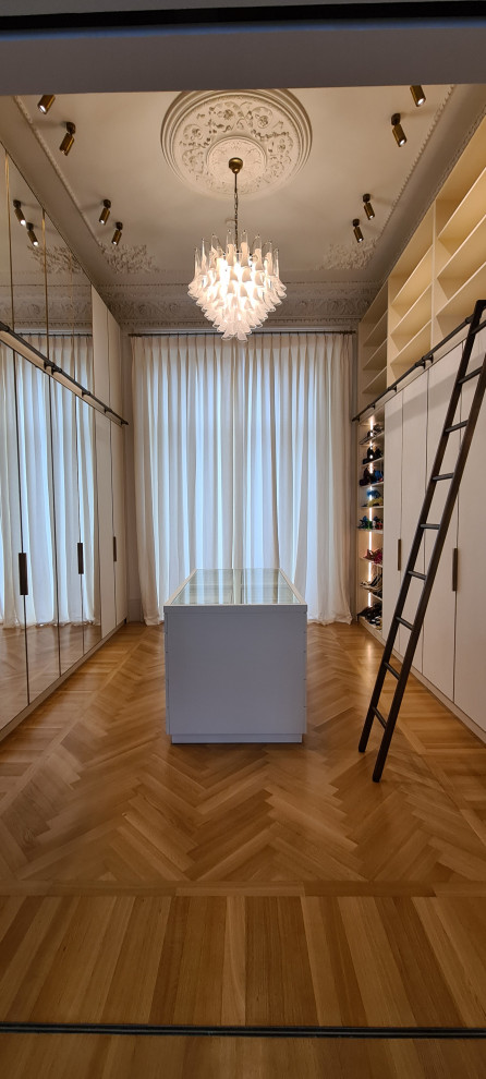 Photo of a large transitional women's storage and wardrobe in London with glass-front cabinets, white cabinets, medium hardwood floors and brown floor.