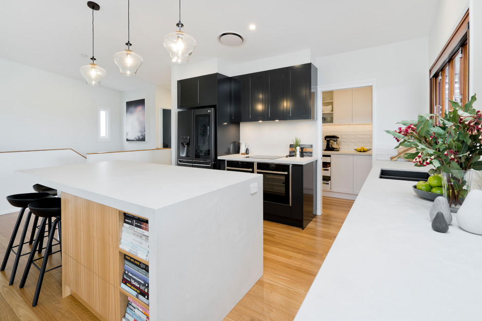 Photo of a large contemporary l-shaped kitchen pantry in Brisbane with an undermount sink, recessed-panel cabinets, black cabinets, quartz benchtops, white splashback, subway tile splashback, black appliances, light hardwood floors, with island, brown floor and white benchtop.
