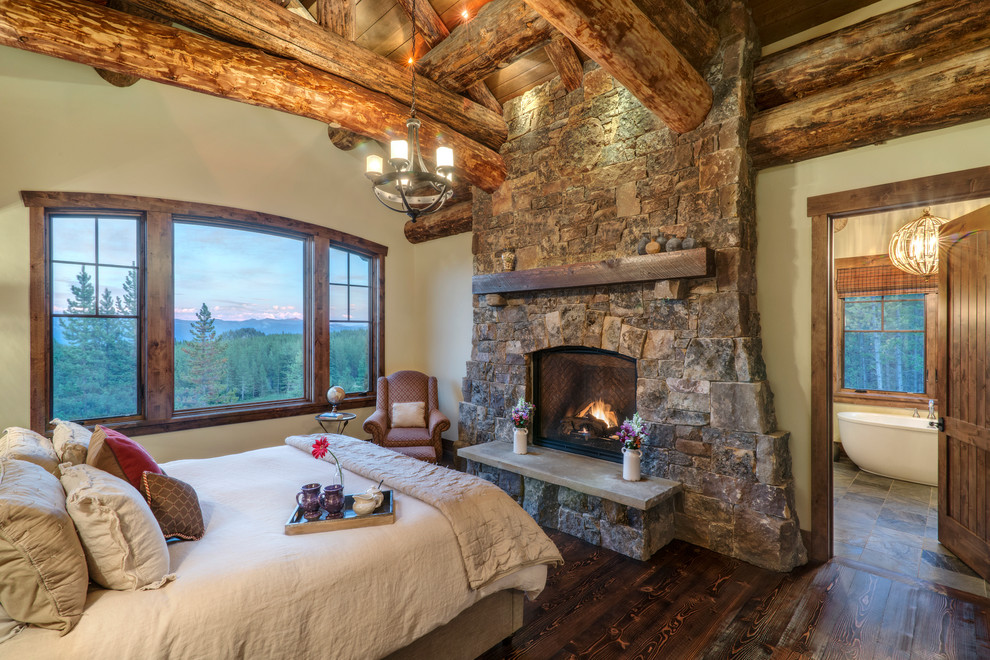 Example of a mountain style brown floor and dark wood floor bedroom design with beige walls, a standard fireplace and a stone fireplace