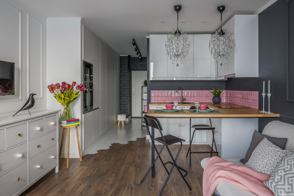 Photo of a small contemporary u-shaped eat-in kitchen in Other with a drop-in sink, flat-panel cabinets, white cabinets, wood benchtops, pink splashback, ceramic splashback, stainless steel appliances, laminate floors, a peninsula, brown floor and brown benchtop.