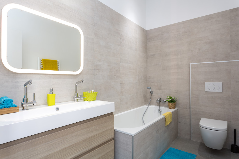 This is an example of a contemporary kids bathroom in Paris with flat-panel cabinets, light wood cabinets, a corner tub, a wall-mount toilet, gray tile, a trough sink and grey floor.