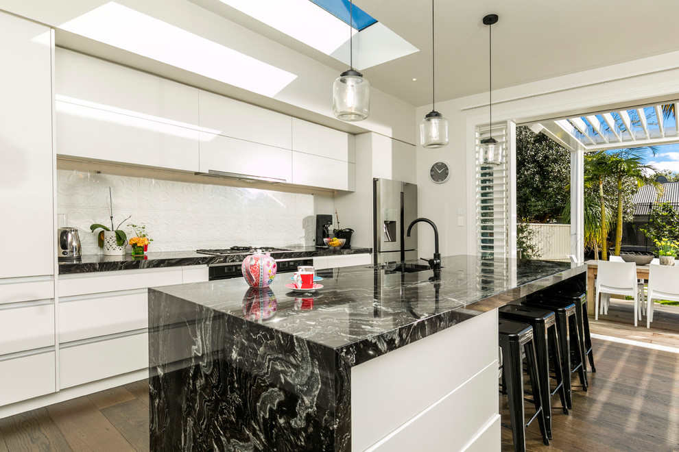 Photo of a mid-sized contemporary l-shaped open plan kitchen in Auckland with an undermount sink, flat-panel cabinets, white cabinets, granite benchtops, white splashback, subway tile splashback, stainless steel appliances, dark hardwood floors and with island.