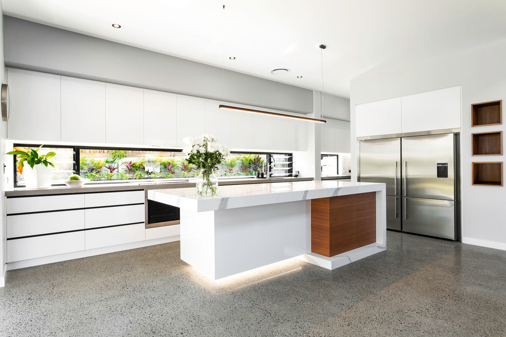 Large contemporary galley open plan kitchen in Brisbane with a drop-in sink, flat-panel cabinets, white cabinets, quartz benchtops, window splashback, stainless steel appliances, concrete floors, with island, grey floor and white benchtop.