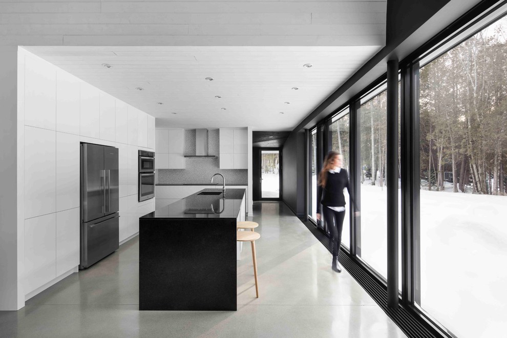 Modern kitchen in Montreal with a single-bowl sink, flat-panel cabinets, white cabinets, grey splashback, stainless steel appliances, concrete floors, with island and grey floor.