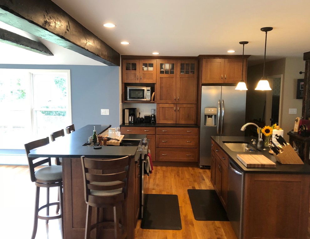 Photo of an arts and crafts galley eat-in kitchen in Portland Maine with an undermount sink, shaker cabinets, brown cabinets, granite benchtops, white splashback, subway tile splashback, stainless steel appliances, light hardwood floors, multiple islands, beige floor and black benchtop.