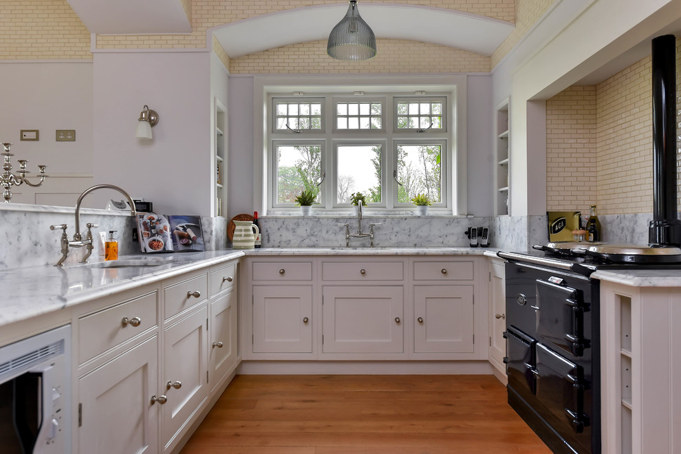 Photo of a traditional u-shaped kitchen in Other with an undermount sink, shaker cabinets, white cabinets, grey splashback, black appliances, medium hardwood floors, a peninsula, brown floor and grey benchtop.