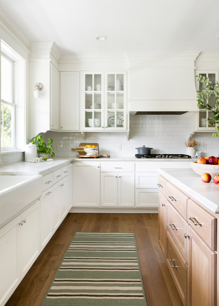 Traditional l-shaped kitchen in Minneapolis with a farmhouse sink, recessed-panel cabinets, medium wood cabinets, white splashback, subway tile splashback, medium hardwood floors, with island, brown floor and white benchtop.
