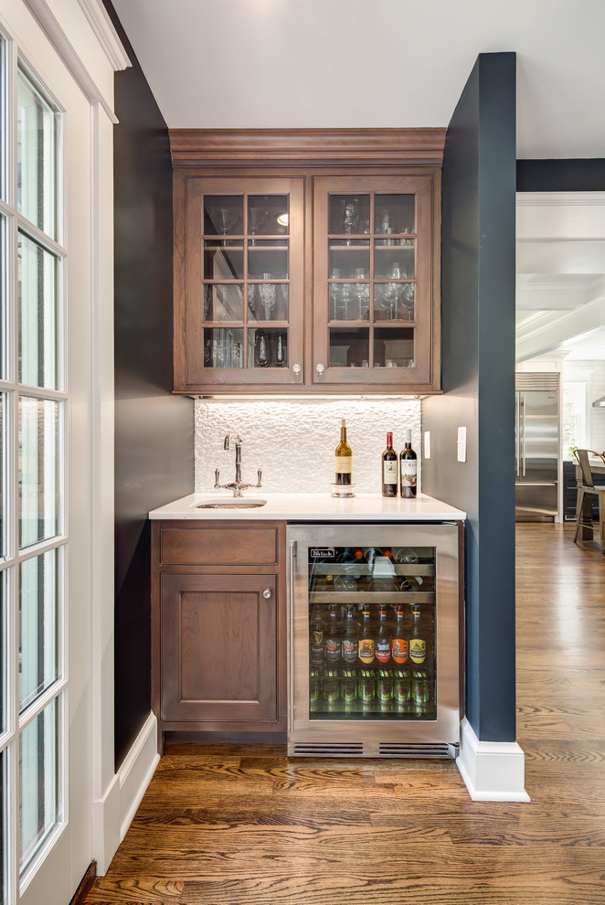 Small transitional single-wall wet bar in DC Metro with an undermount sink, glass-front cabinets, medium wood cabinets, quartz benchtops, white splashback, stone tile splashback, medium hardwood floors, brown floor and white benchtop.