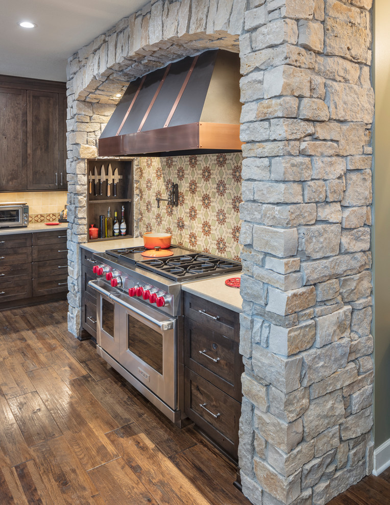 This is an example of a mid-sized country open plan kitchen in Kansas City with a farmhouse sink, shaker cabinets, distressed cabinets, quartz benchtops, multi-coloured splashback, terra-cotta splashback, stainless steel appliances, medium hardwood floors, with island and brown floor.