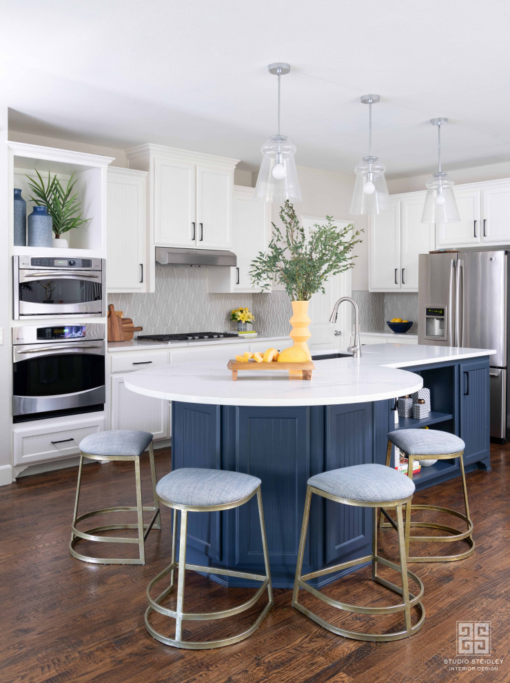 This is an example of a mid-sized transitional l-shaped eat-in kitchen in Dallas with a drop-in sink, recessed-panel cabinets, blue cabinets, marble benchtops, grey splashback, porcelain splashback, stainless steel appliances, medium hardwood floors, with island, brown floor and white benchtop.