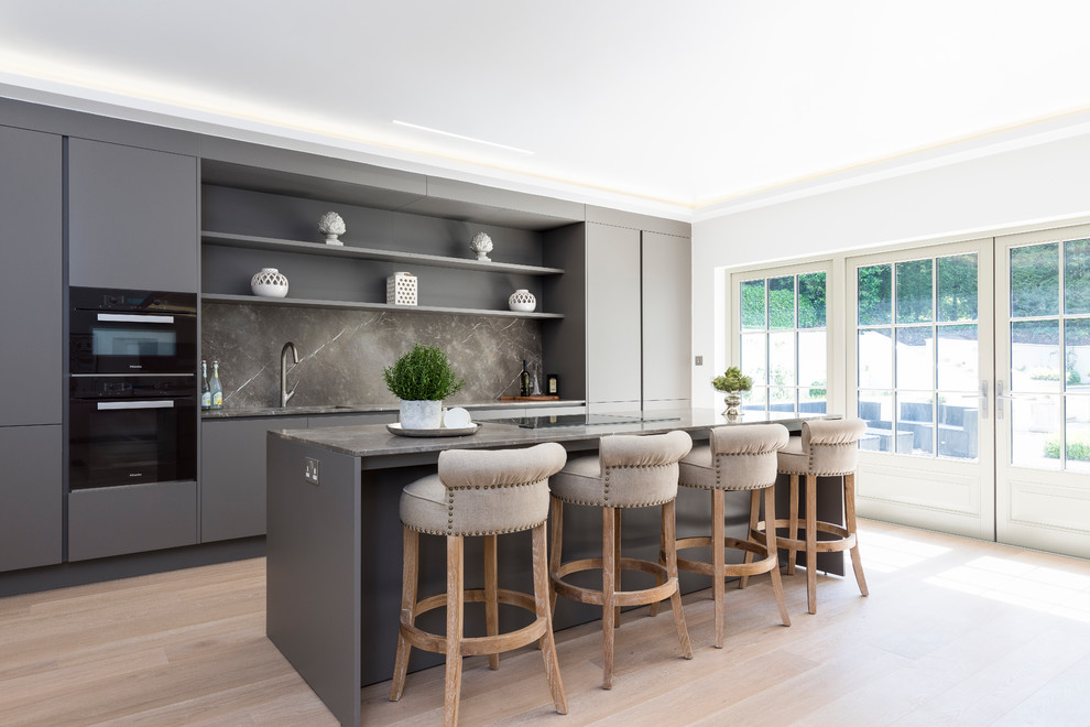 This is an example of a large transitional single-wall kitchen in London with flat-panel cabinets, black cabinets, marble benchtops, black splashback, marble splashback, black appliances, light hardwood floors, a peninsula and beige floor.