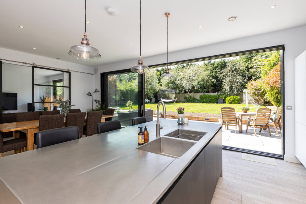 Large contemporary l-shaped open plan kitchen in Hertfordshire with flat-panel cabinets, white cabinets, stainless steel worktops, black appliances, an island and feature lighting.