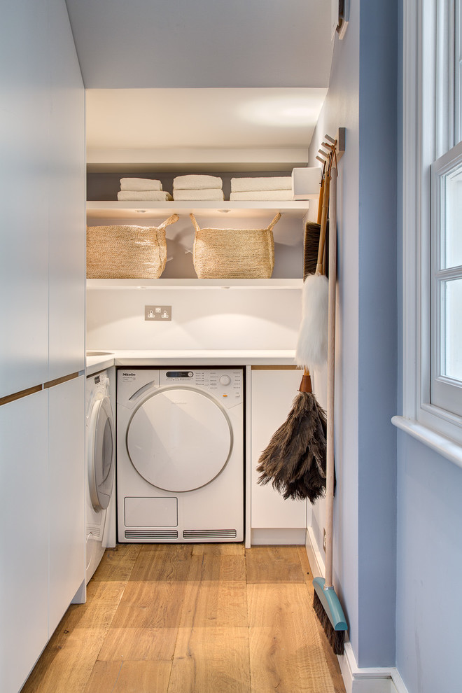 Inspiration for a contemporary l-shaped laundry room in London with flat-panel cabinets, white walls, medium hardwood floors, a side-by-side washer and dryer and white cabinets.