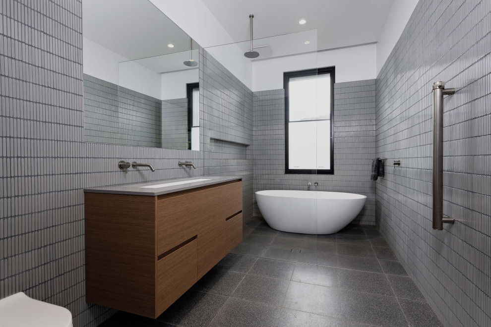 This is an example of a medium sized modern ensuite bathroom in Adelaide with recessed-panel cabinets, medium wood cabinets, a freestanding bath, grey tiles, matchstick tiles, grey walls, ceramic flooring, an integrated sink, solid surface worktops, grey floors, white worktops, a single sink, a built in vanity unit and wood walls.