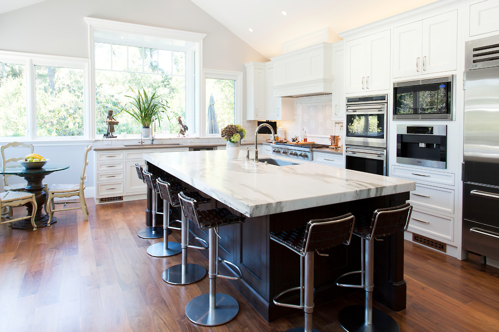 Photo of a transitional l-shaped open plan kitchen in San Francisco with an undermount sink, recessed-panel cabinets, white cabinets, white splashback, white appliances, medium hardwood floors, with island and brown floor.