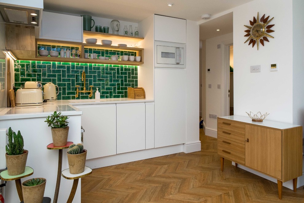 Photo of a contemporary l-shaped kitchen in London with flat-panel cabinets, white cabinets, green splashback, white appliances, medium hardwood floors, no island and brown floor.