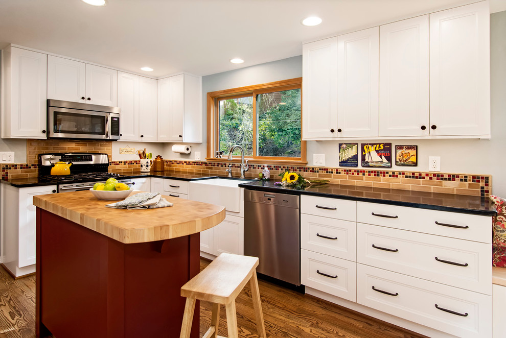 Photo of a mid-sized country l-shaped kitchen in Seattle with a farmhouse sink, shaker cabinets, white cabinets, solid surface benchtops, brown splashback, subway tile splashback, stainless steel appliances, medium hardwood floors and with island.