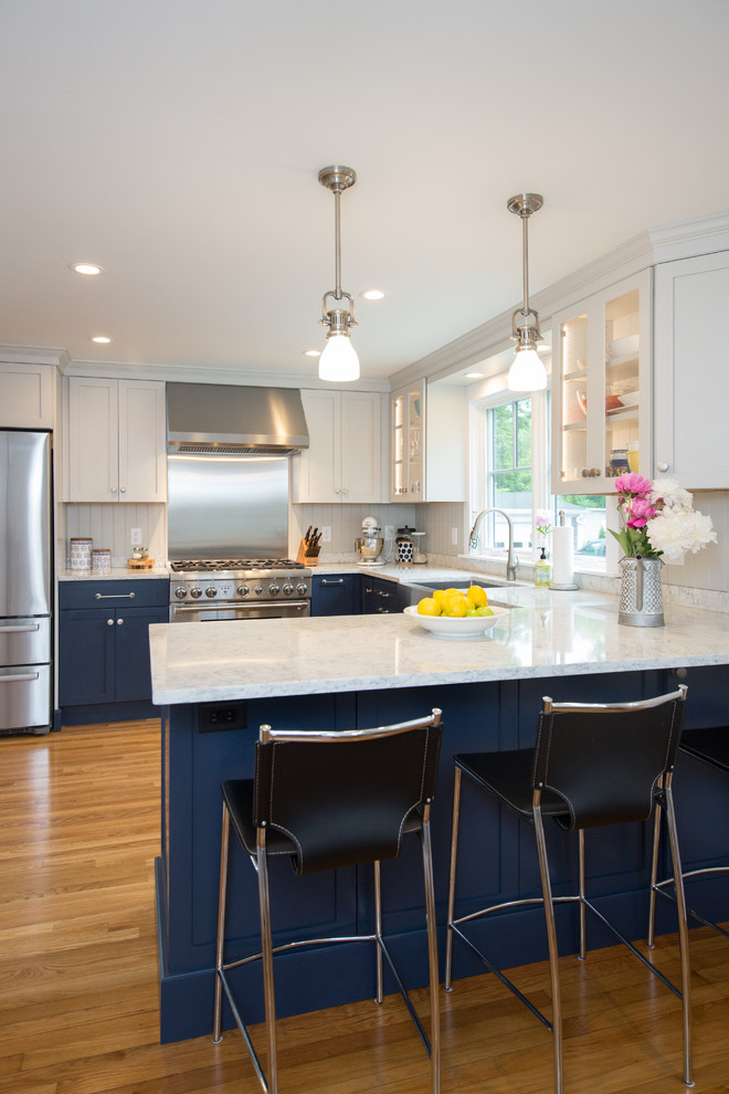 This is an example of a beach style eat-in kitchen in Boston with a farmhouse sink, shaker cabinets, blue cabinets, quartz benchtops, grey splashback, timber splashback, stainless steel appliances, medium hardwood floors, a peninsula and white benchtop.