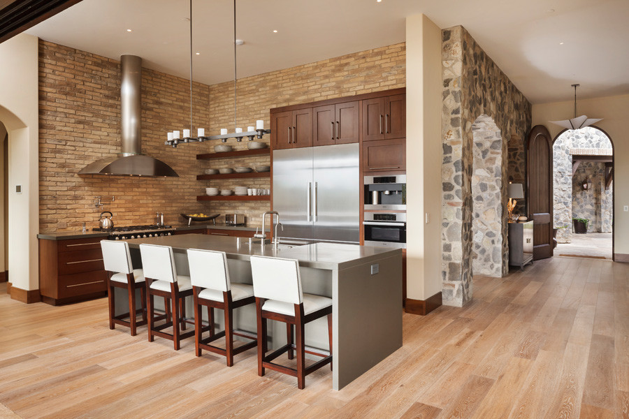 Inspiration for a large industrial l-shaped open plan kitchen in San Francisco with shaker cabinets, dark wood cabinets, quartz benchtops, beige splashback, brick splashback, stainless steel appliances, light hardwood floors, with island and a farmhouse sink.