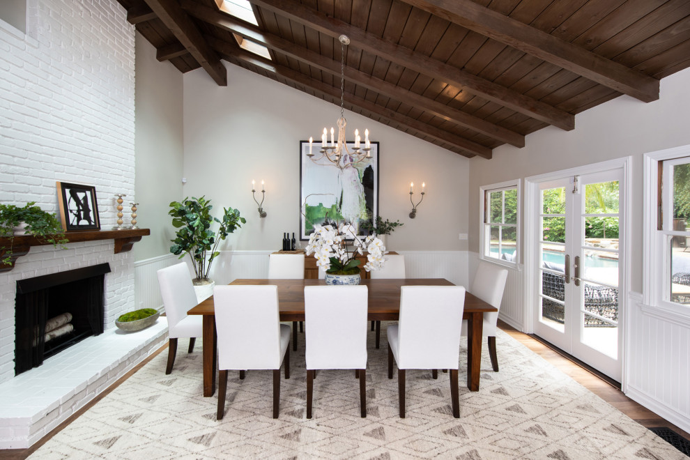 This is an example of a large beach style dining room in Los Angeles with grey walls, dark hardwood floors, a two-sided fireplace, a brick fireplace surround, brown floor, exposed beam and decorative wall panelling.