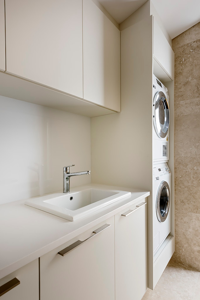 Inspiration for a mid-sized modern galley dedicated laundry room in Perth with an utility sink, flat-panel cabinets, beige cabinets, quartz benchtops, white splashback, engineered quartz splashback, beige walls, travertine floors, a stacked washer and dryer, beige floor, white benchtop and vaulted.