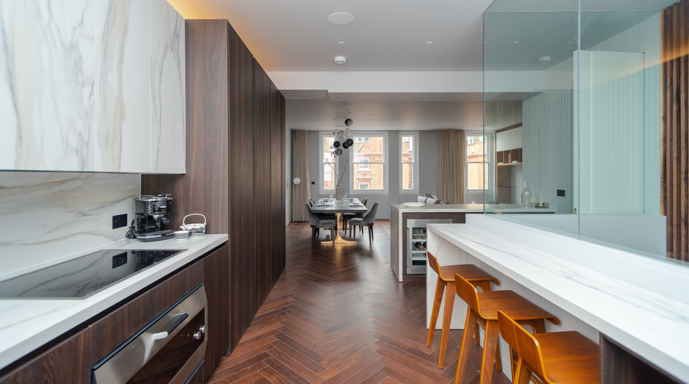 Design ideas for a contemporary galley kitchen in London with flat-panel cabinets, dark wood cabinets, white splashback, brown floor, white benchtop, stainless steel appliances, dark hardwood floors and with island.