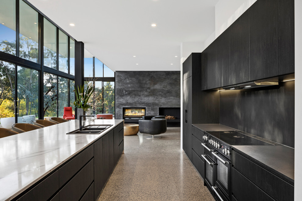 Design ideas for a large modern galley open plan kitchen in Melbourne with a drop-in sink, flat-panel cabinets, black cabinets, quartz benchtops, black splashback, engineered quartz splashback, black appliances, concrete floors, with island, multi-coloured benchtop and vaulted.