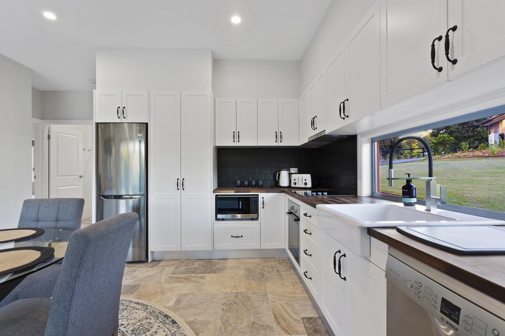 Inspiration for a small modern kitchen in Sunshine Coast with a farmhouse sink, shaker cabinets, white cabinets, wood benchtops, black splashback, subway tile splashback, stainless steel appliances, travertine floors and multi-coloured floor.