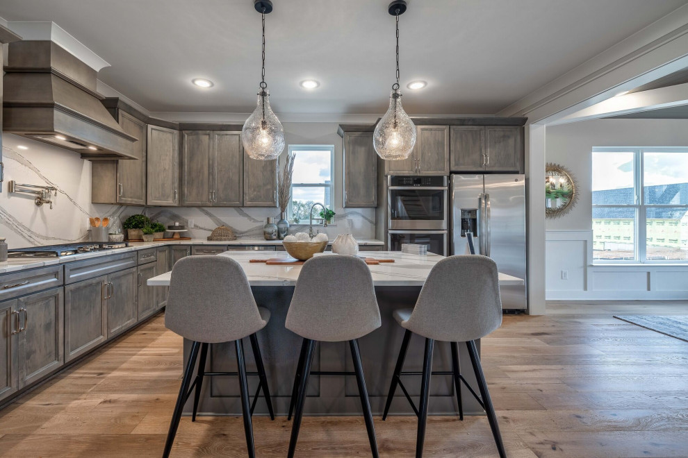 Large rustic l-shaped open plan kitchen in Nashville with recessed-panel cabinets, grey cabinets, engineered stone countertops, white splashback, engineered quartz splashback, stainless steel appliances, light hardwood flooring, an island and white worktops.