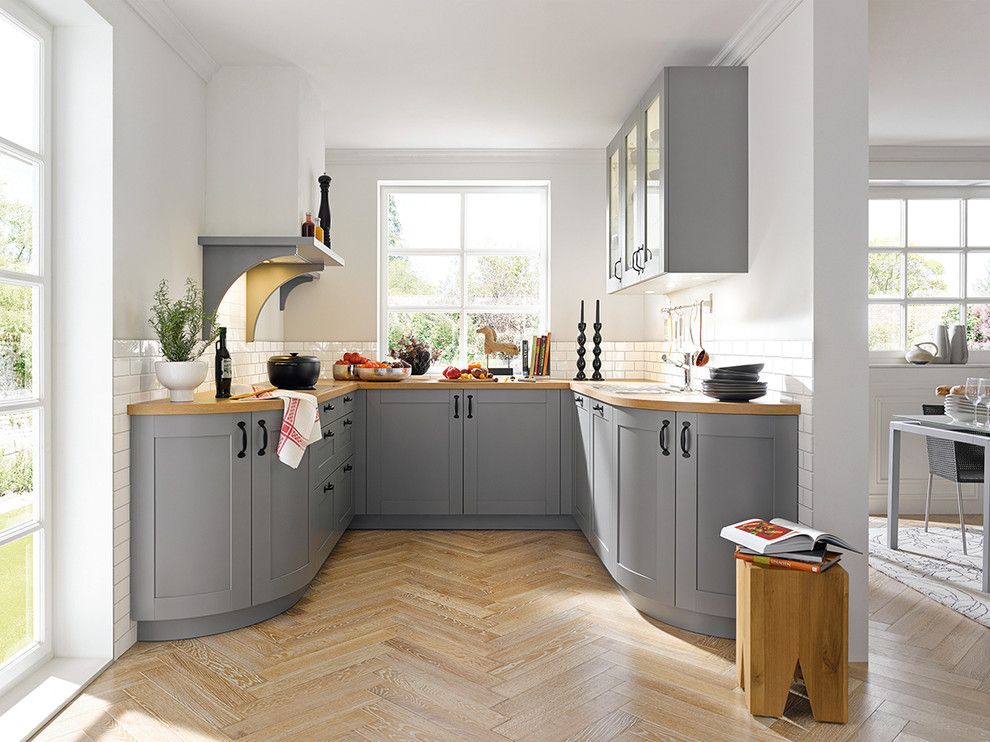 This is an example of a mid-sized country u-shaped open plan kitchen in Other with raised-panel cabinets, grey cabinets, wood benchtops, white splashback, ceramic splashback, medium hardwood floors and no island.