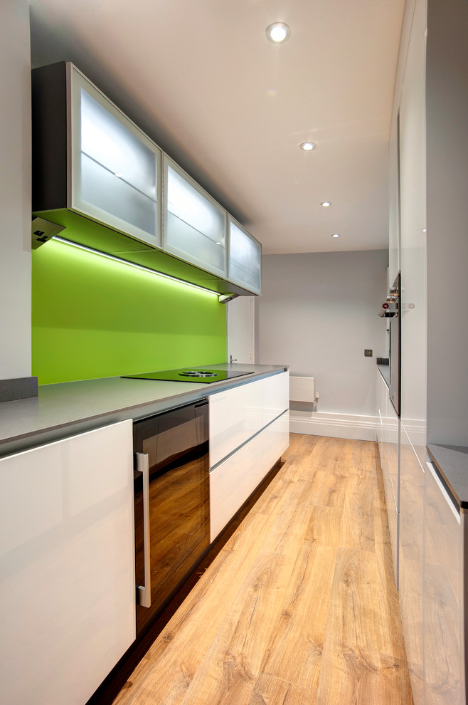 Photo of a small modern galley separate kitchen in Other with flat-panel cabinets, white cabinets, quartz benchtops, green splashback, glass sheet splashback, black appliances, laminate floors, no island and grey benchtop.
